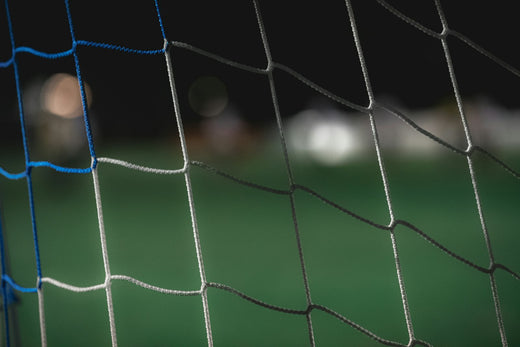 Close-up of a white and blue football goal net with a blurred green pitch behind it