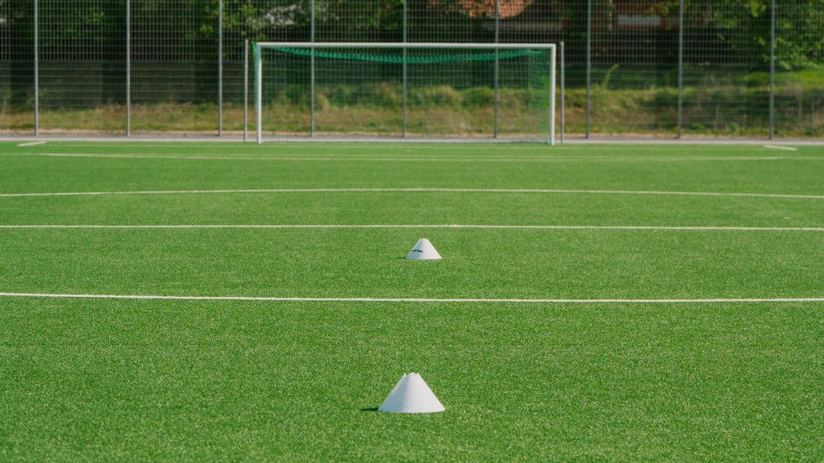 Two white training cones in a line on an artificial football pitch, with an empty goal and a mesh fence behind